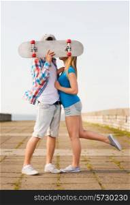 holidays, vacation, love and people concept - couple kissing and hiding their faces behind skateboard outdoors