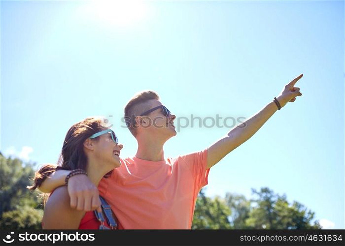 holidays, vacation, love and people concept - happy smiling teenage couple hugging and pointing finger up to something at summer