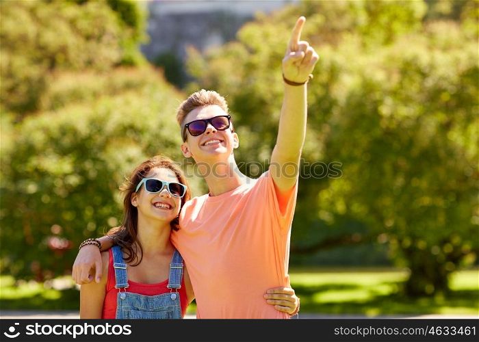 holidays, vacation, love and people concept - happy smiling teenage couple hugging and pointing finger to something at summer park