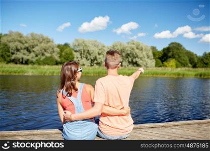 holidays, vacation, love and people concept - happy smiling teenage couple hugging and pointing finger to something on river berth at summer