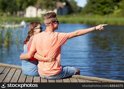 holidays, vacation, love and people concept - happy smiling teenage couple hugging and pointing finger to something on river berth at summer