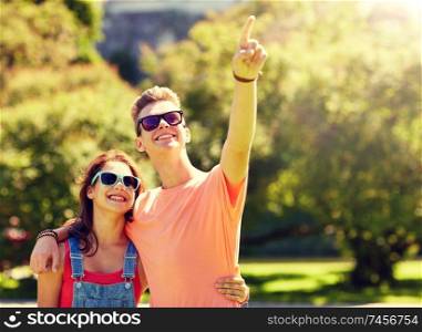 holidays, vacation, love and people concept - happy smiling teenage couple hugging and pointing finger to something at summer park. happy couple pointing finger at summer park