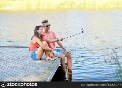 holidays, vacation, love and people concept - happy teenage couple taking picture by smartphone selfie stick and sitting on river berth at summer