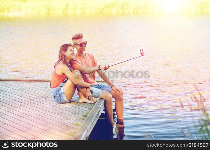 holidays, vacation, love and people concept - happy teenage couple taking picture by smartphone selfie stick and sitting on river berth at summer. happy teenage couple taking selfie on smartphone