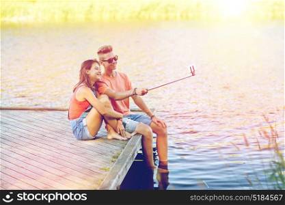 holidays, vacation, love and people concept - happy teenage couple taking picture by smartphone selfie stick and sitting on river berth at summer. happy teenage couple taking selfie on smartphone