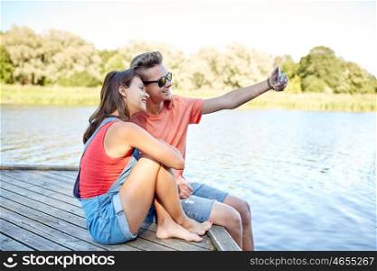 holidays, vacation, love and people concept - happy teenage couple taking selfie on smartphone and sitting on river berth at summer