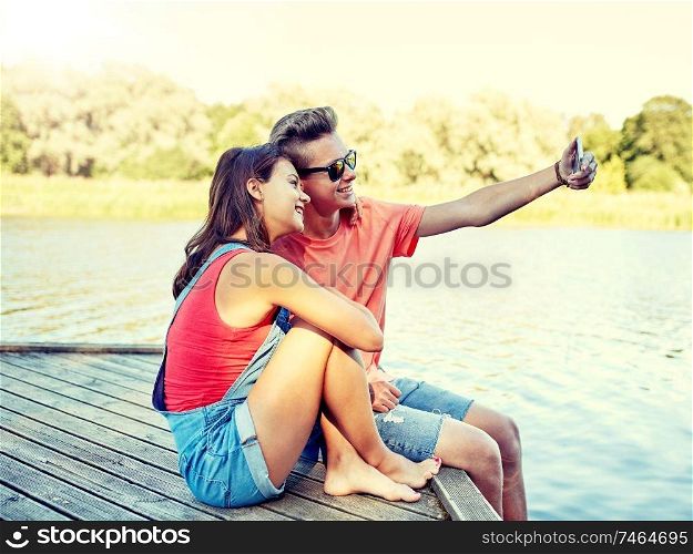 holidays, vacation, love and people concept - happy teenage couple taking selfie on smartphone and sitting on river berth at summer. happy teenage couple taking selfie on smartphone