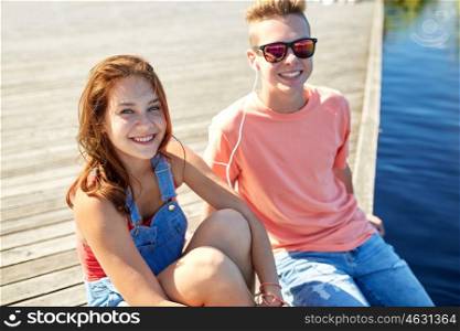 holidays, vacation, love and people concept - happy teenage couple with earphones sitting on river berth and listening to music at summer