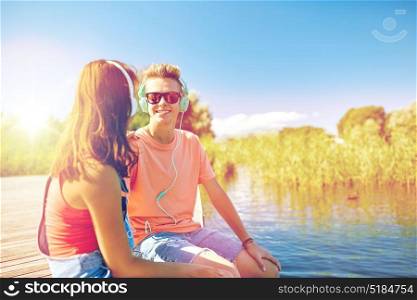 holidays, vacation, love and people concept - happy teenage couple with earphones sitting on river berth and listening to music at summer. happy teenage couple with earphones on river berth