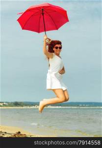 Holidays, vacation travel and freedom concept. Beautiful redhaired happy girl jumping with red umbrella on beach.