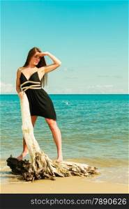 Holidays, vacation travel and freedom concept. Full length of beautiful sexy girl long hair on beach. Young pretty tanned woman in summer dress relaxing on the sea coast standing on dry tree root