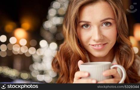 holidays, winter and people concept - close up of happy young woman with cup of coffee or tea over christmas tree lights background. close up of happy woman with coffee at christmas