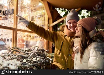 holidays, winter and people concept - happy couple at christmas market shop window in evening choosing souvenirs. happy couple at christmas market shop window