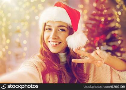 holidays, winter and people concept - happy young woman in santa hat taking selfie over christmas tree at home. happy woman taking selfie over christmas tree