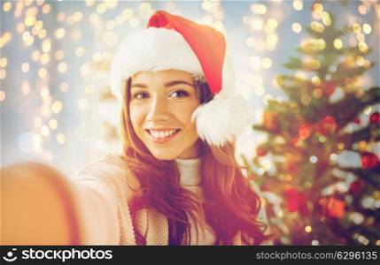 holidays, winter and people concept - happy young woman or teenage girl in santa hat over christmas tree at home. happy woman in santa hat over christmas tree