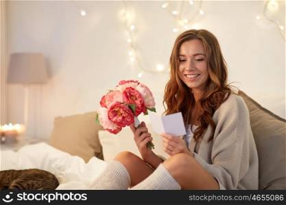holidays, winter, birthday and people concept - happy young woman with flowers reading greeting card in bed at home bedroom