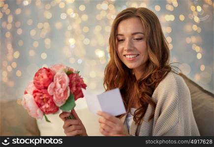 holidays, winter, birthday and people concept - happy young woman with flowers reading greeting card in bed at home bedroom. happy woman with flowers and greeting card at home
