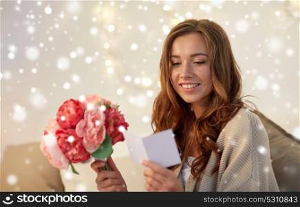 holidays, winter, birthday and people concept - happy young woman with flowers reading greeting card in bed at home over snow. happy woman with flowers and greeting card at home