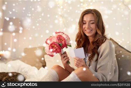 holidays, winter, birthday and people concept - happy young woman with flowers reading greeting card in bed at home bedroom over snow. happy woman with flowers and greeting card at home