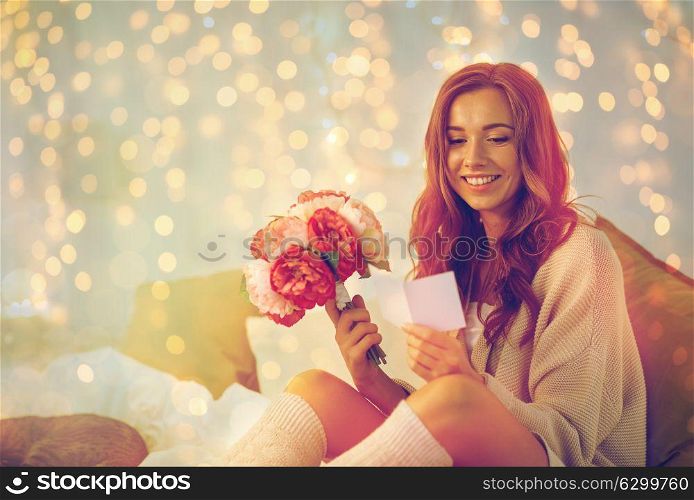 holidays, winter, birthday and people concept - happy young woman with flowers reading greeting card in bed at home bedroom. happy woman with flowers and greeting card at home
