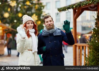 holidays, winter, christmas, gesture and people concept - happy couple of tourists in warm clothes waving hands in old town