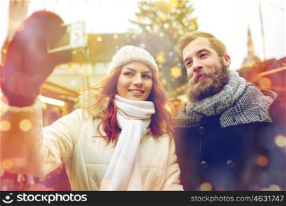 holidays, winter, christmas, technology and people concept - happy couple of tourists in warm clothes taking selfie with smartphone in old town