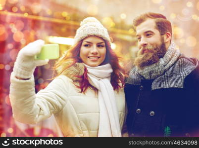 holidays, winter, christmas, technology and people concept - happy couple of tourists in warm clothes taking selfie with smartphone in old town