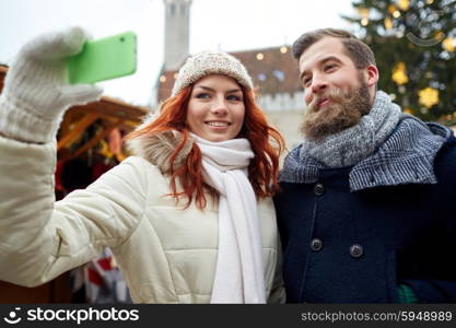 holidays, winter, christmas, technology and people concept - happy couple of tourists in warm clothes taking selfie with smartphone in old town