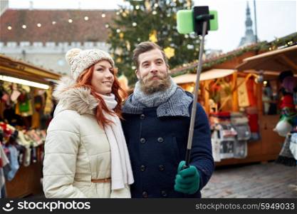 holidays, winter, christmas, technology and people concept - happy couple of tourists in warm clothes taking picture with smartphone selfie stick in old town