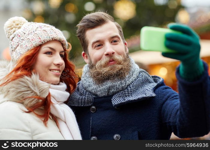 holidays, winter, christmas, technology and people concept - happy couple of tourists in warm clothes taking selfie with smartphone in old town