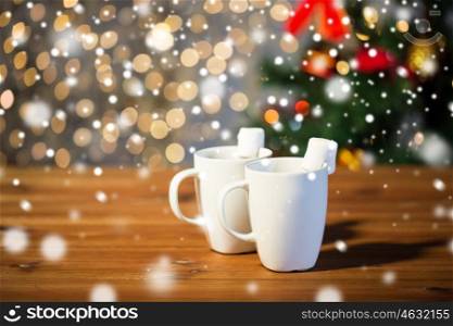 holidays, winter, food and drinks concept - close up of cups with hot chocolate or cocoa drinks and marshmallow on wooden table over lights