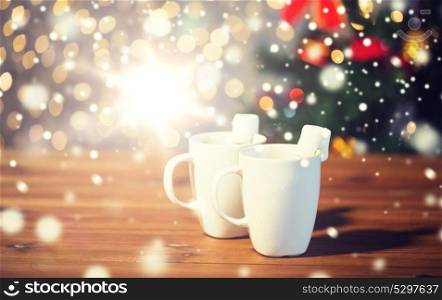 holidays, winter, food and drinks concept - close up of cups with hot chocolate or cocoa drinks and marshmallow on wooden table over lights. cups of hot chocolate with marshmallow on wood