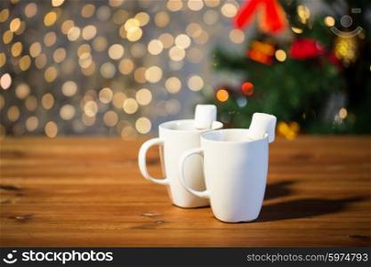 holidays, winter, food and drinks concept - close up of cups with hot chocolate or cocoa drinks and marshmallow on wooden table over lights