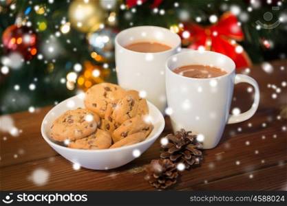 holidays, winter, food and drinks concept - close up of oatmeal cookies, cups with hot chocolate or cocoa drink and pinecones on wooden table over christmas tree background