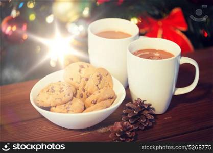 holidays, winter, food and drinks concept - close up of oatmeal cookies, cups with hot chocolate or cocoa drink and pinecones on wooden table over christmas tree background. oat cookies and hot chocolate over christmas tree