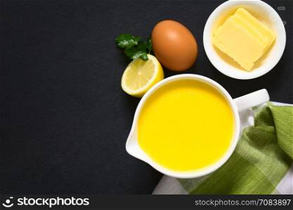 Hollandaise sauce, a basic sauce of the French cuisine, served in a sauce boat with ingredients (egg, butter, lemon, pepper) on the side, photographed overhead on slate with natural light