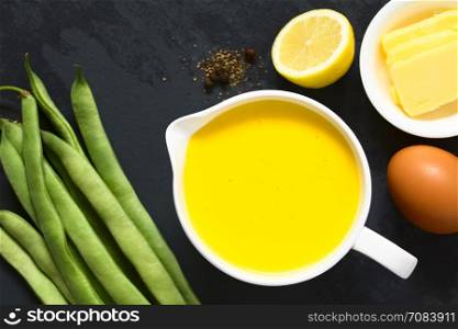 Hollandaise sauce, a basic sauce of the French cuisine, served in a sauce boat with ingredients (egg, butter, lemon, pepper) and raw green beans on the side, photographed overhead on slate with natural light (Selective Focus, Focus on the sauce)