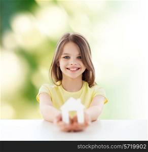 home, education, nature, childhood and people concept - beautiful little girl sitting at table holding white house cutout over green background