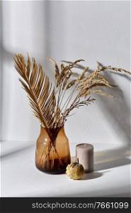 home improvement and decoration concept - still life of decorative dried flowers in brown glass vase, candle and pumpkin dropping shadows on white surface. dried flowers in glass vase candle and pumpkin