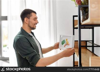 home improvement, decoration and people concept - happy smiling man placing watercolor picture in frame to shelf. man decorating home with picture in frame
