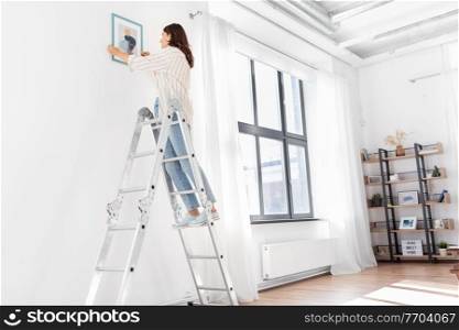 home improvement, decoration and people concept - happy smiling woman on ladder decorating home with art. woman on ladder decorating home with art