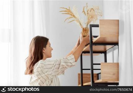home improvement, decoration and people concept - happy smiling woman placing dried flowers or herbs in vase to shelf. woman decorating home with dried flowers in vase