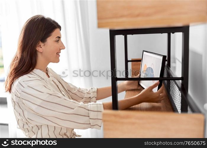 home improvement, decoration and people concept - happy smiling woman placing picture in frame to shelf. woman decorating home with picture in frame