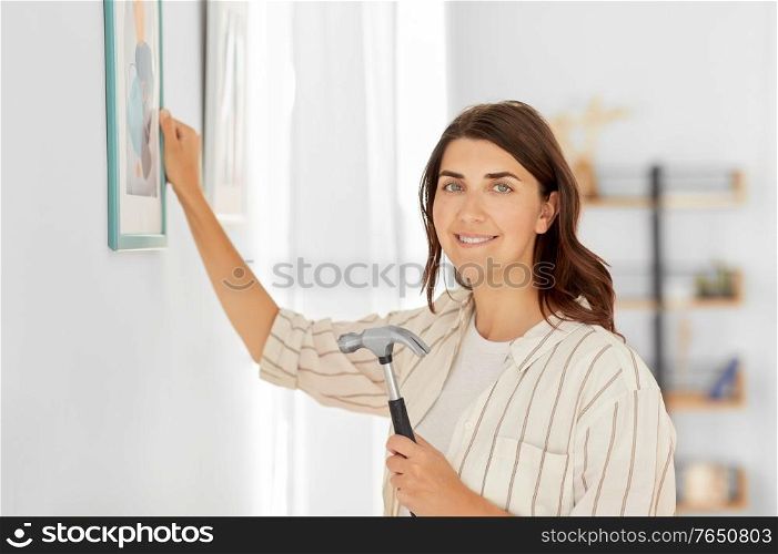 home improvement, decoration and people concept - happy smiling woman with hammer hanging picture in frame on wall. woman decorating home with picture in frame