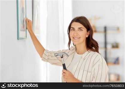 home improvement, decoration and people concept - happy smiling woman with hammer hanging picture in frame on wall. woman decorating home with picture in frame