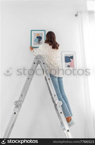 home improvement, decoration and people concept - woman on ladder hanging picture in frame on wall. woman on ladder hanging picture in frame on wall