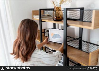 home improvement, decoration and people concept - woman placing picture in frame to shelf. woman decorating home with picture in frame