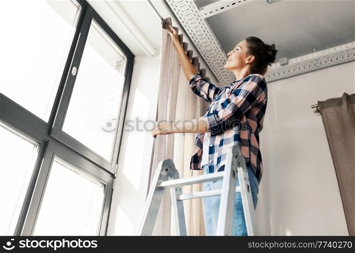 home improvement, decoration and renovation concept - happy smiling woman on ladder hanging curtains. woman on ladder hanging curtains at home