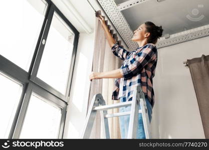 home improvement, decoration and renovation concept - happy smiling woman on ladder hanging curtains. woman on ladder hanging curtains at home