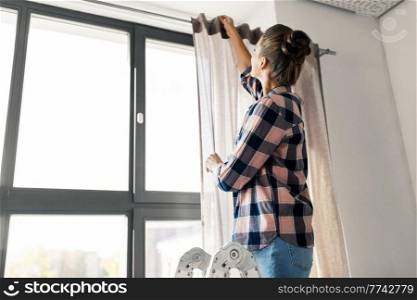 home improvement, decoration and renovation concept - happy woman on ladder hanging curtains. woman on ladder hanging curtains at home